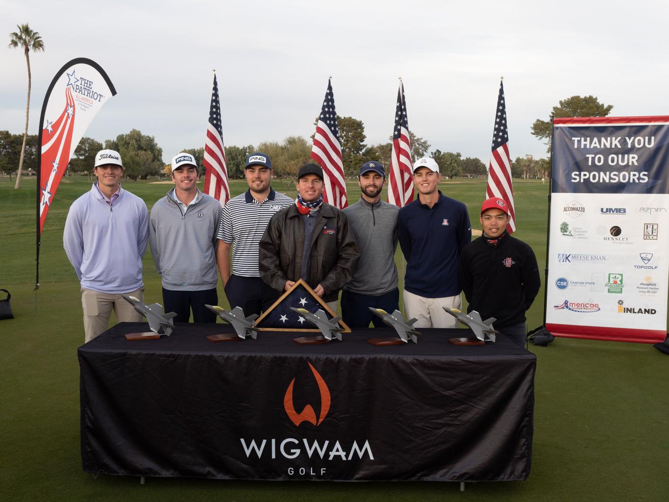 Photo of 7 of the 8 top 5 finishers of the Patriot All-America stadning behind a table with trophies and American flags in the background.